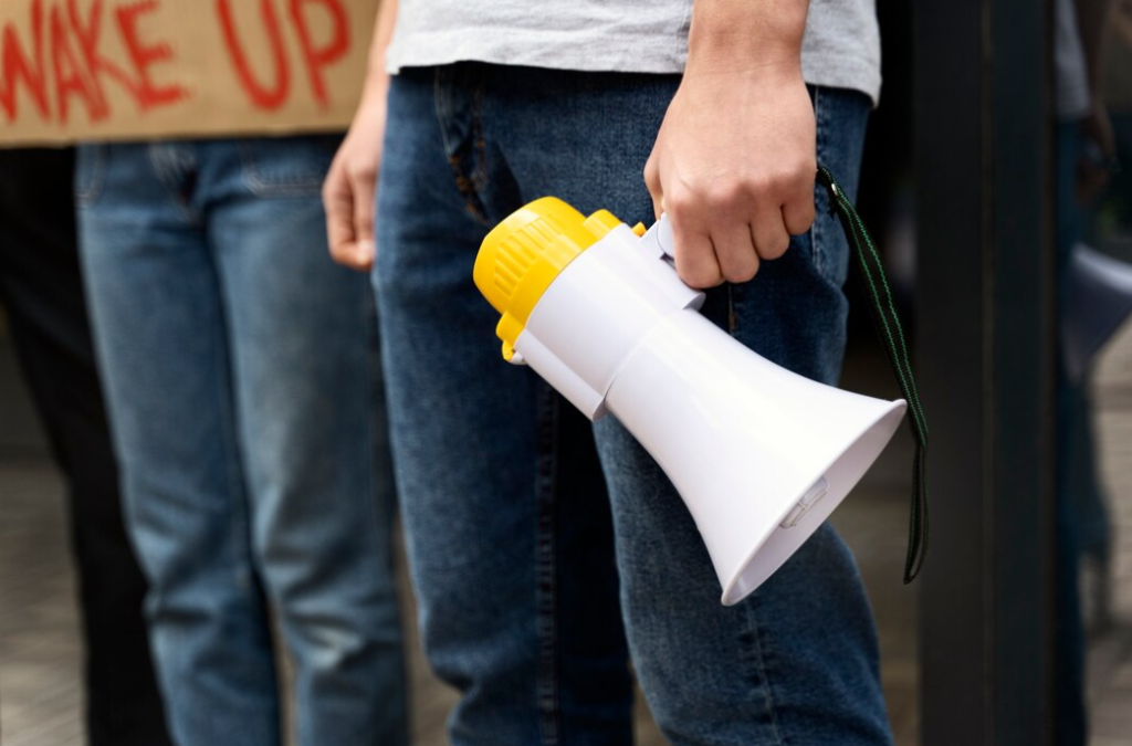 A hand grips a megaphone near a "WAKE UP" sign during a protest